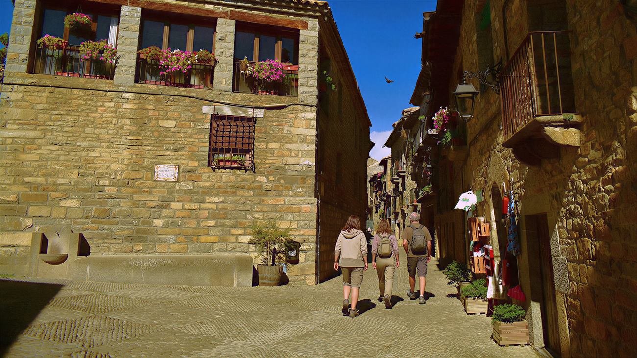 Visitantes paseando por una calle del casco antiguo de Aínsa, con balcones con flores